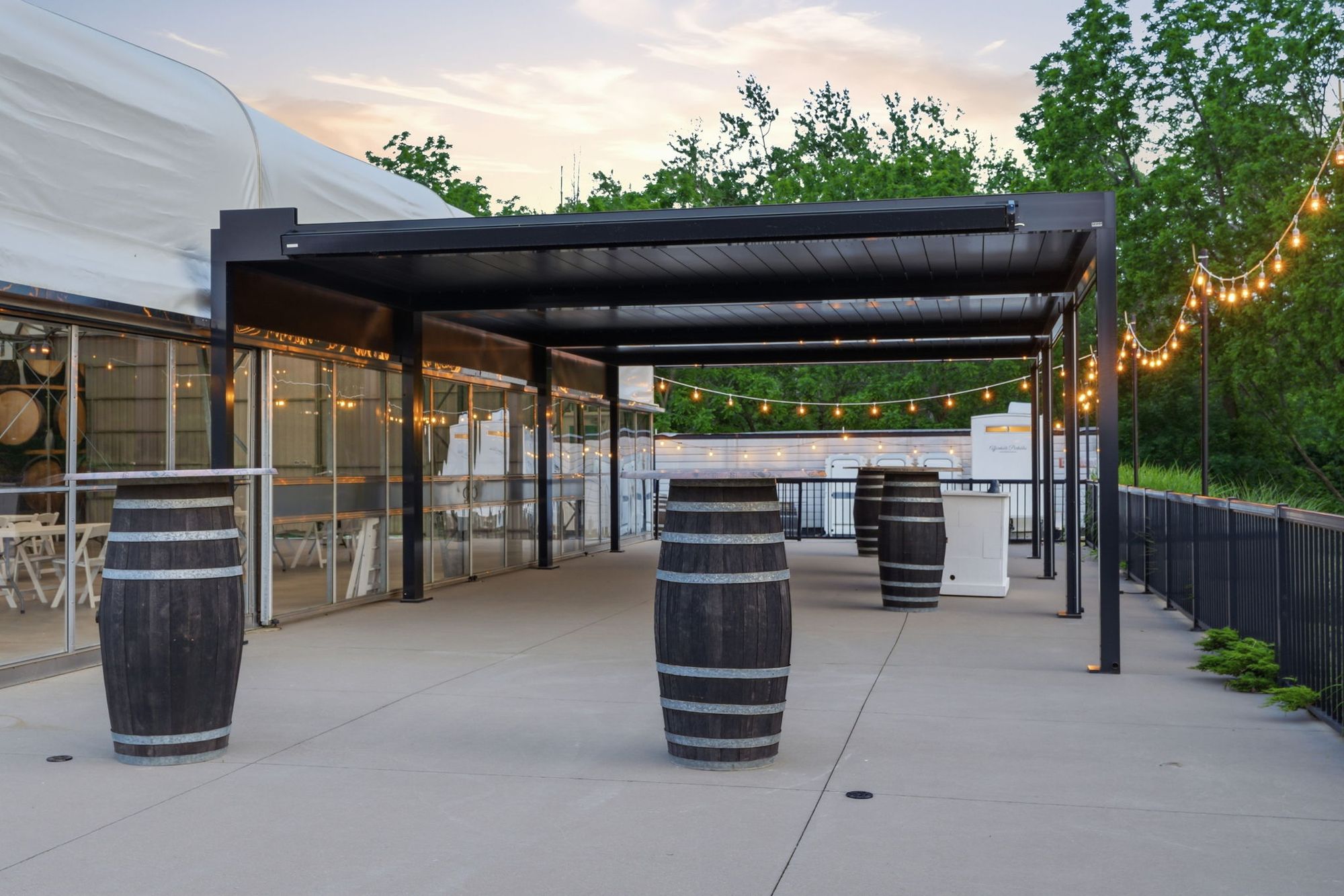 The event space's covered patio at dusk, barrel tables under string lights